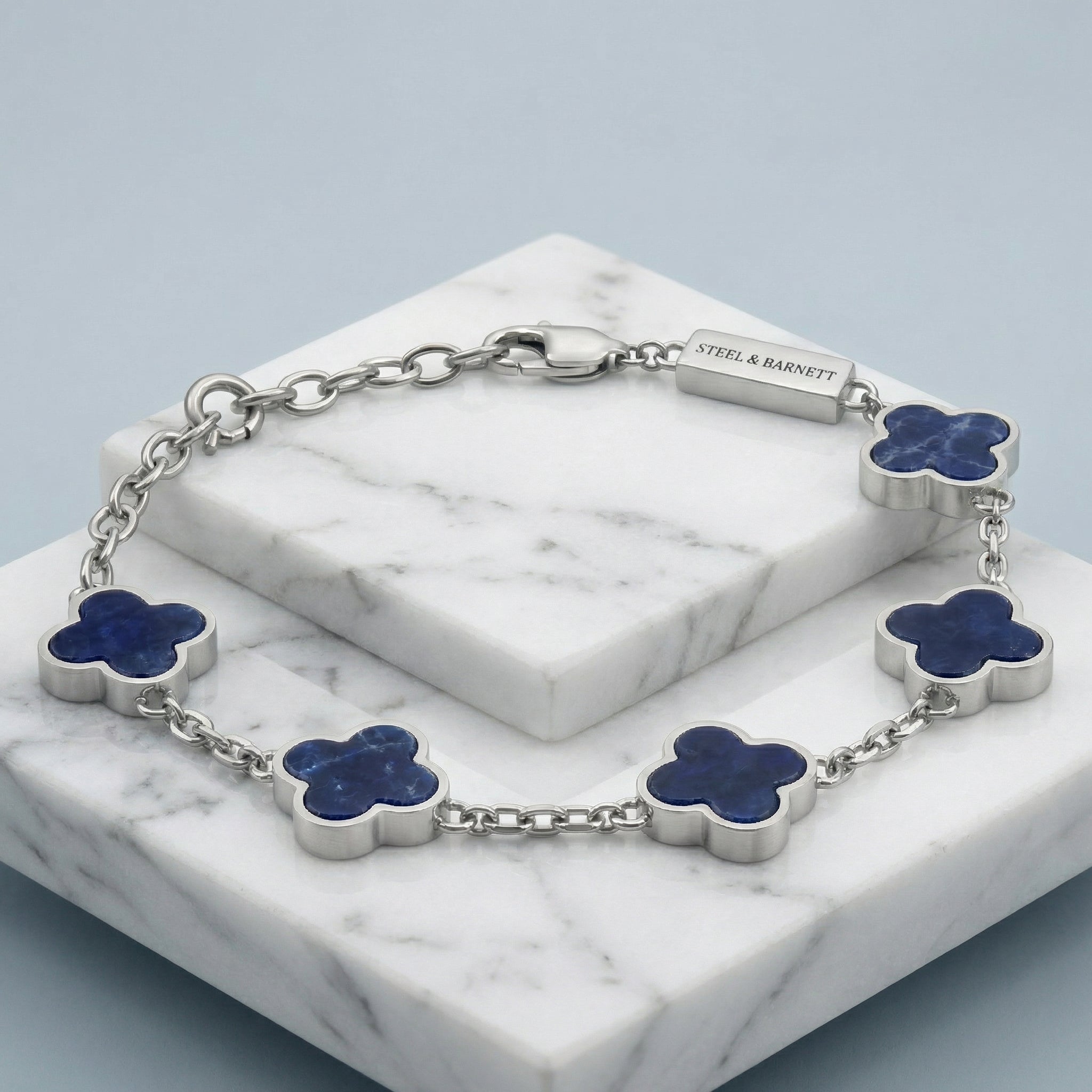 Silver toned men’s bracelet with blue clover shaped stone inlays in stainless steel chain displayed on marble blocks