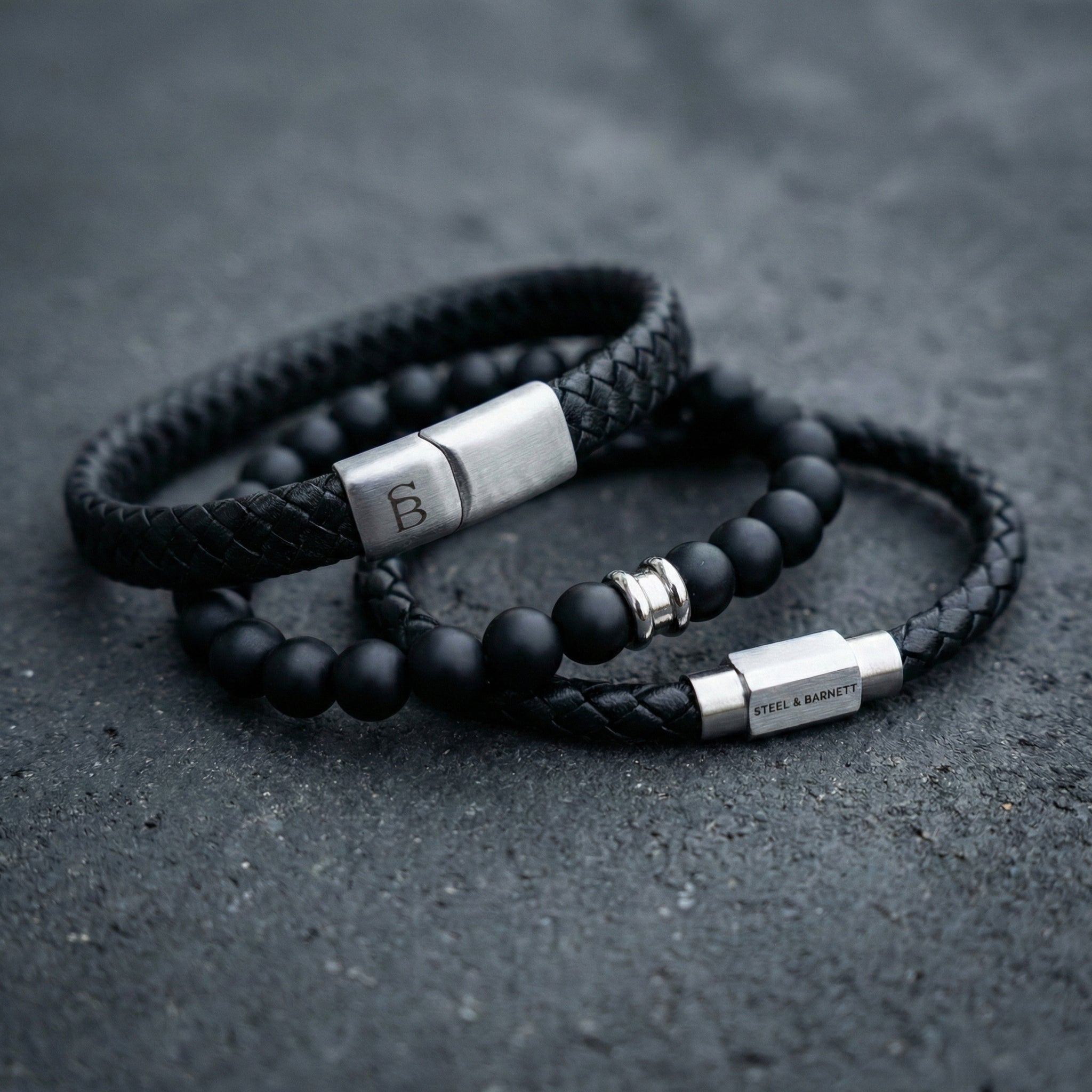Black braided leather men’s bracelet set with matte black bead bracelet and brushed stainless steel clasp detail on dark stone surface