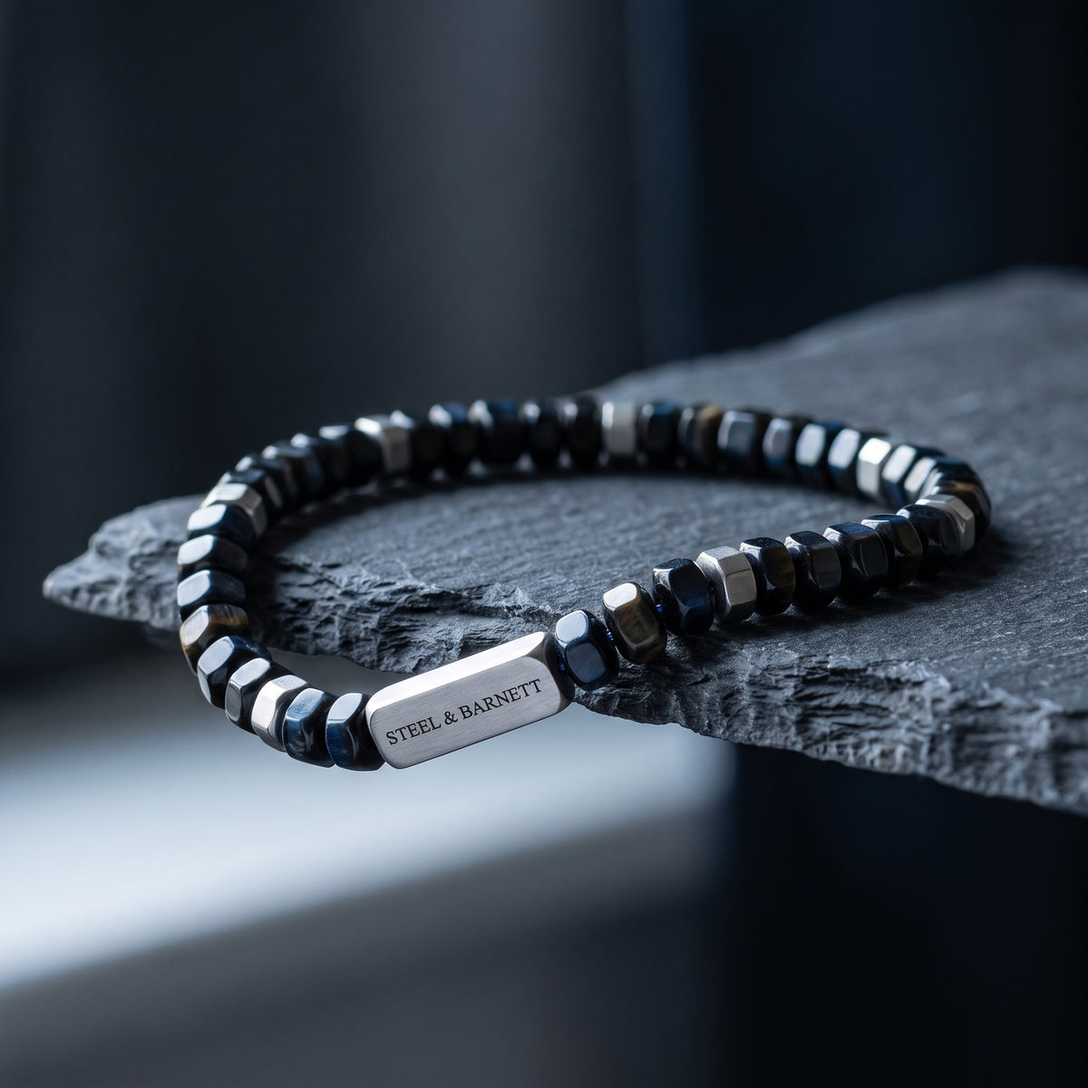 A beaded stretch bracelet with faceted black and silver beads, featuring a polished silver rectangular plate engraved with 'STEEL & BARNETT', resting on a dark textured stone slab.