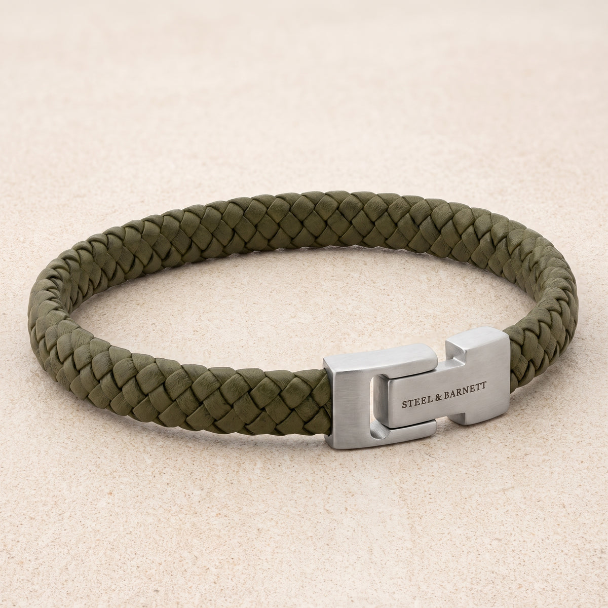 An olive green braided leather bracelet with a silver-toned metal clasp engraved with 'STEEL & BARNETT', displayed on a light beige textured background.