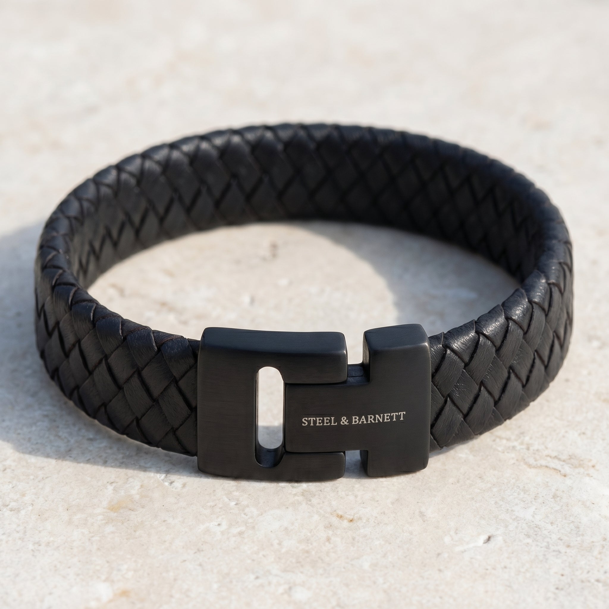 Black braided leather men's bracelet with matte black stainless steel magnetic clasp on a light stone surface close up