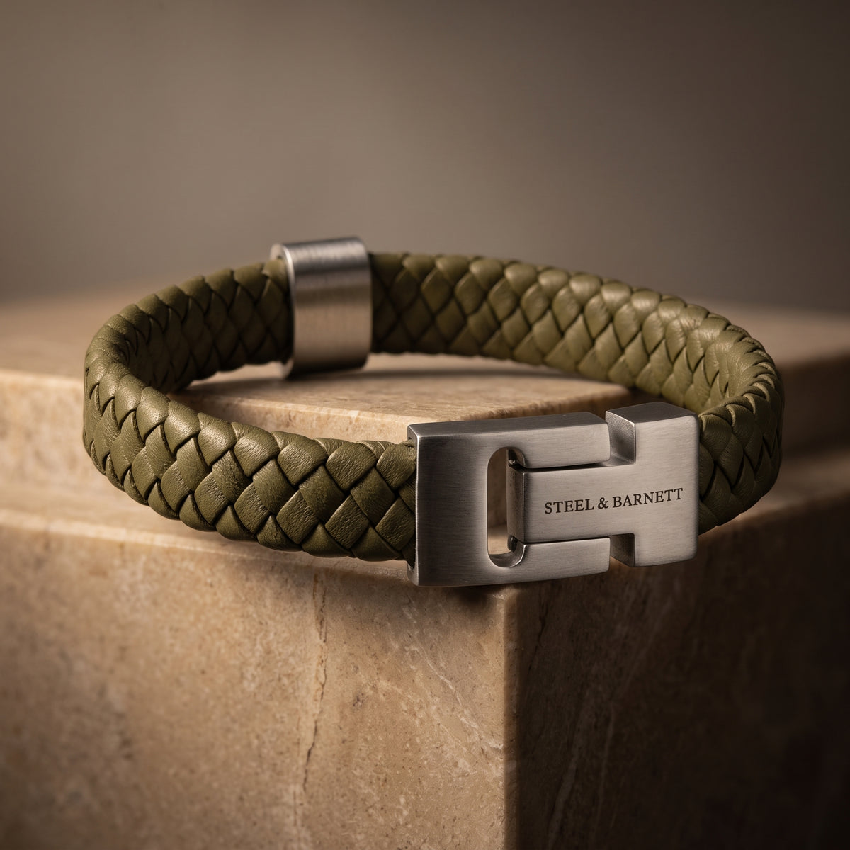 Olive green braided leather men's bracelet with a brushed stainless steel clasp displayed on a stone surface