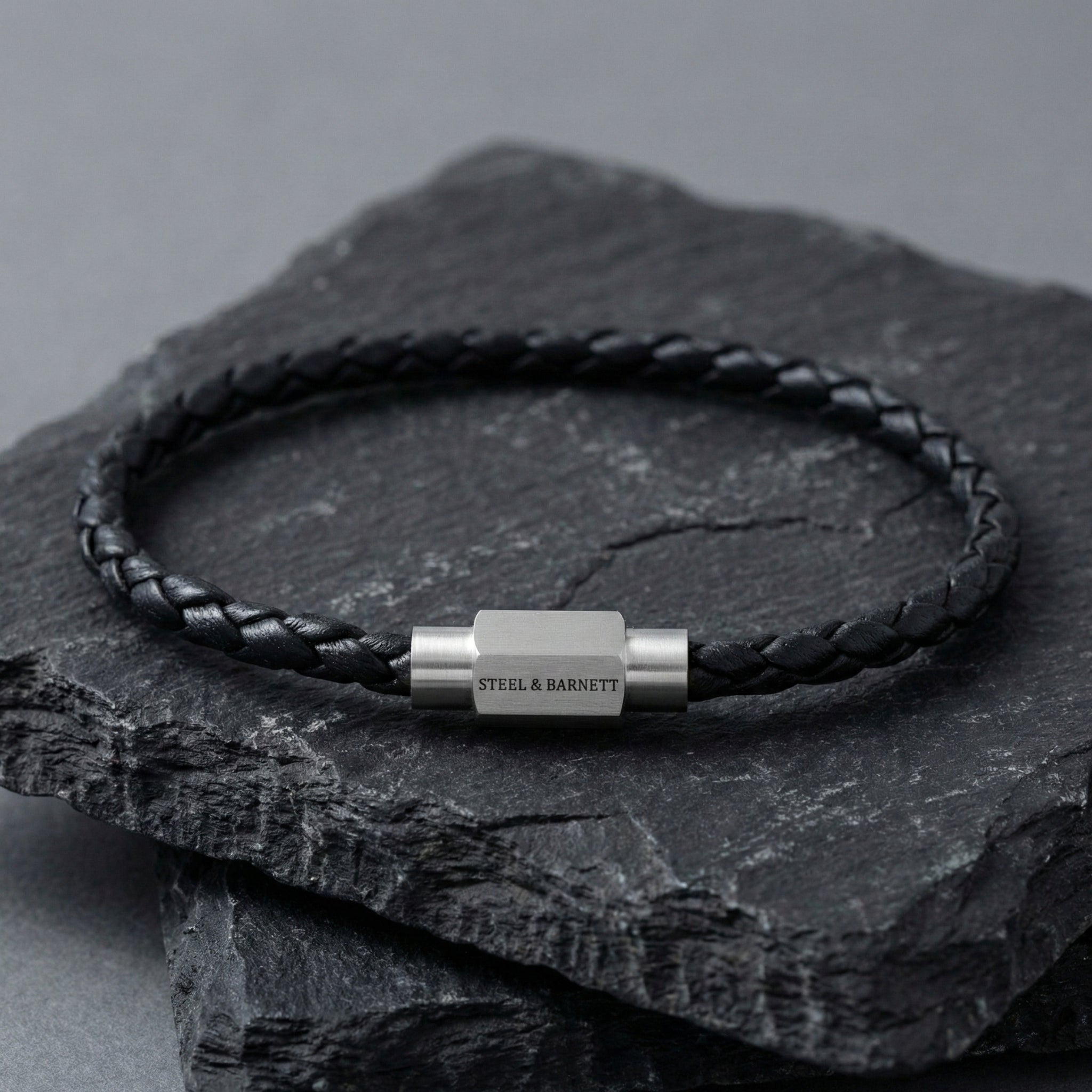 Black braided leather men’s bracelet with brushed stainless steel magnetic clasp on a dark stone surface
