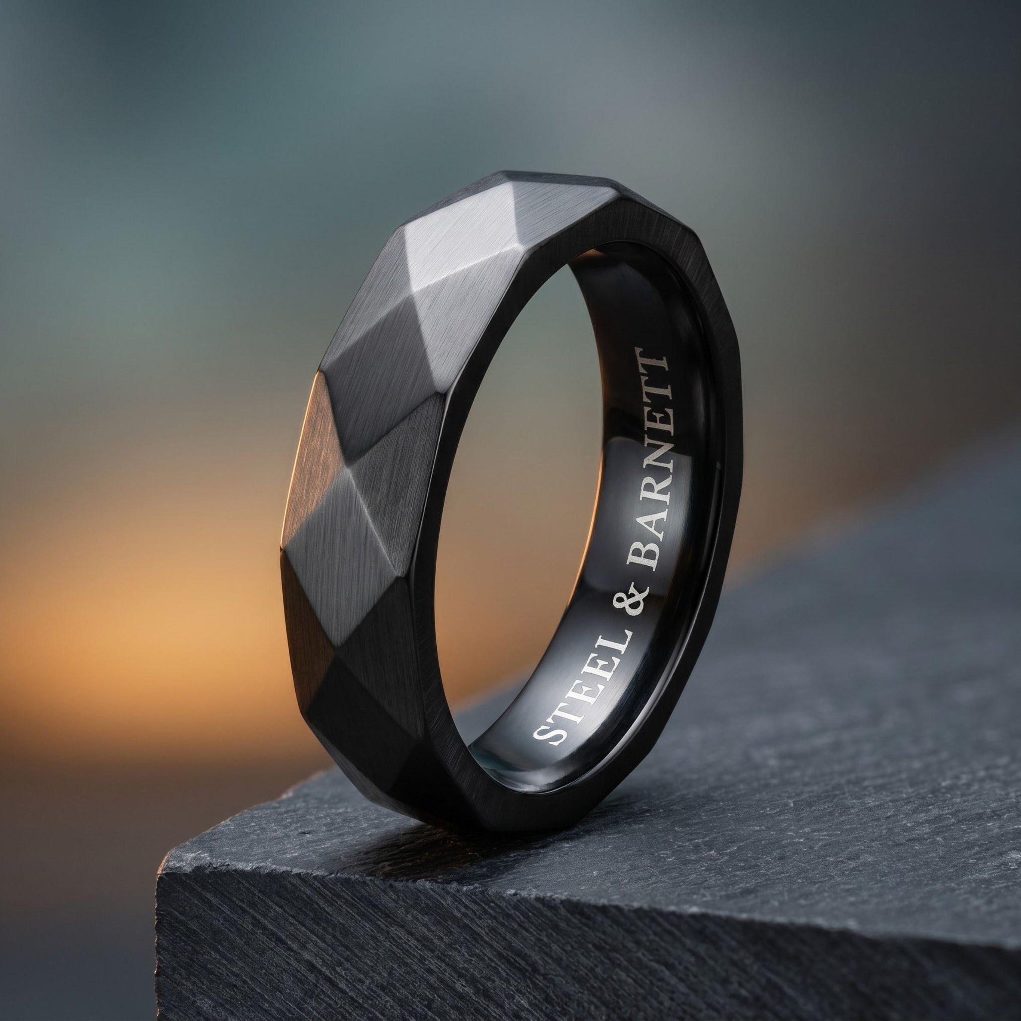 Black stainless steel men's ring with a brushed faceted geometric design displayed upright on a dark stone surface