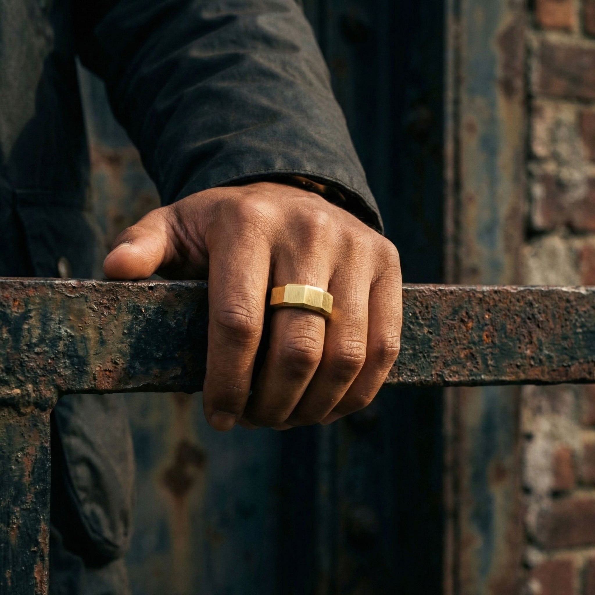 Gold stainless steel men's ring with a brushed geometric design worn on a hand resting on a weathered industrial steel railing
