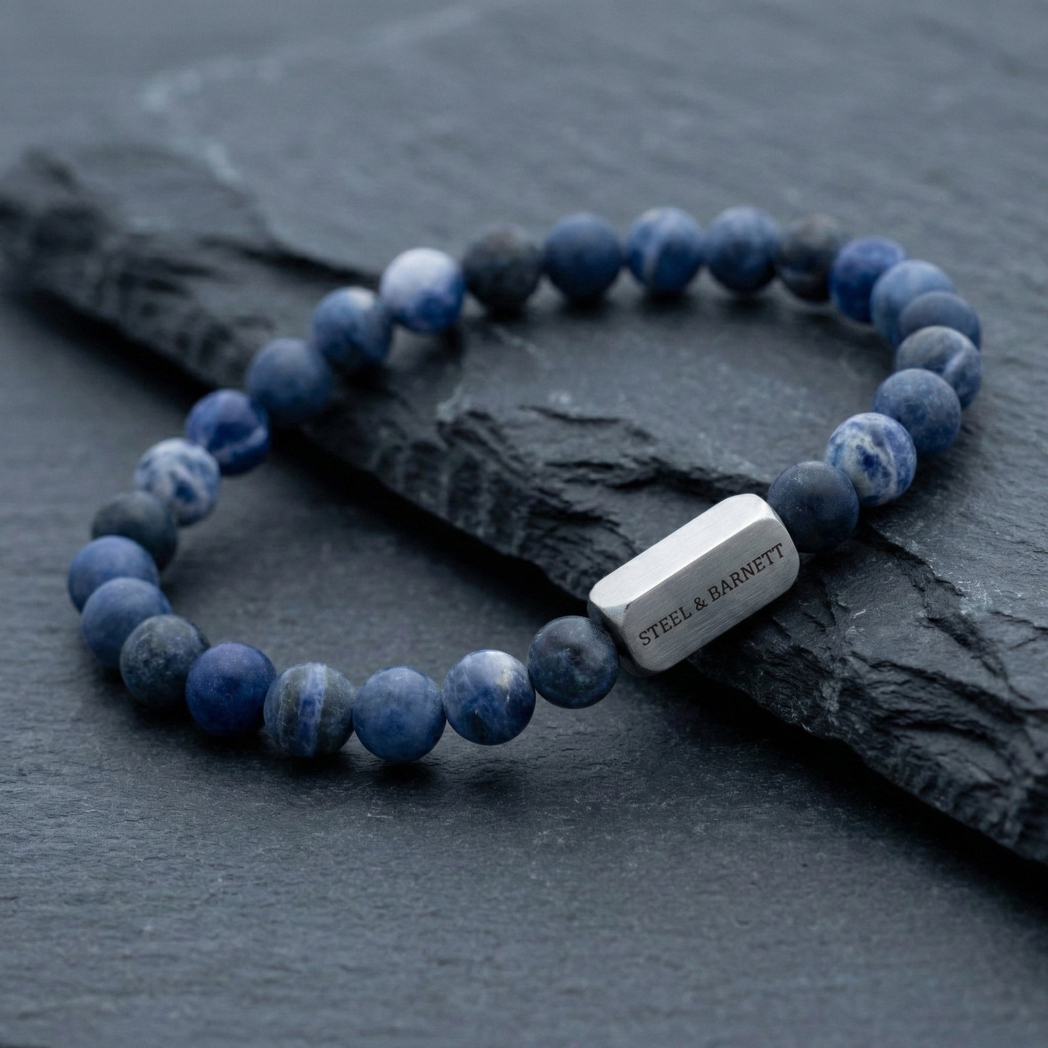 Blue sodalite men’s bracelet with round natural stone beads and brushed stainless steel bar on dark stone surface