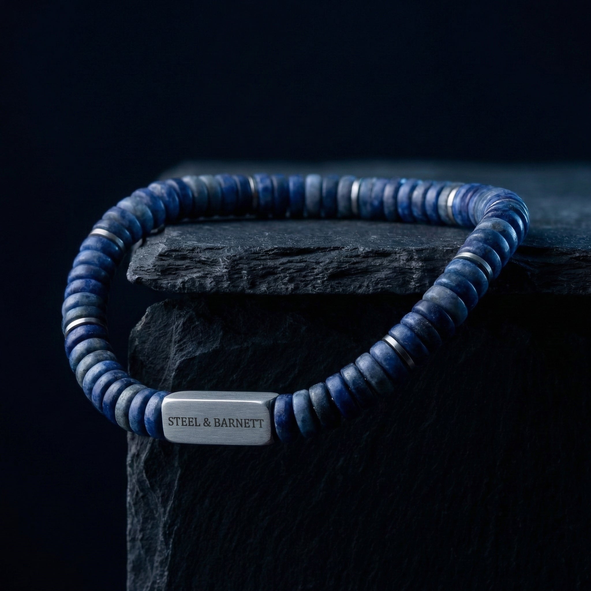 Blue natural stone men's beaded bracelet with brushed stainless steel accent plate displayed on a dark stone block close up detail