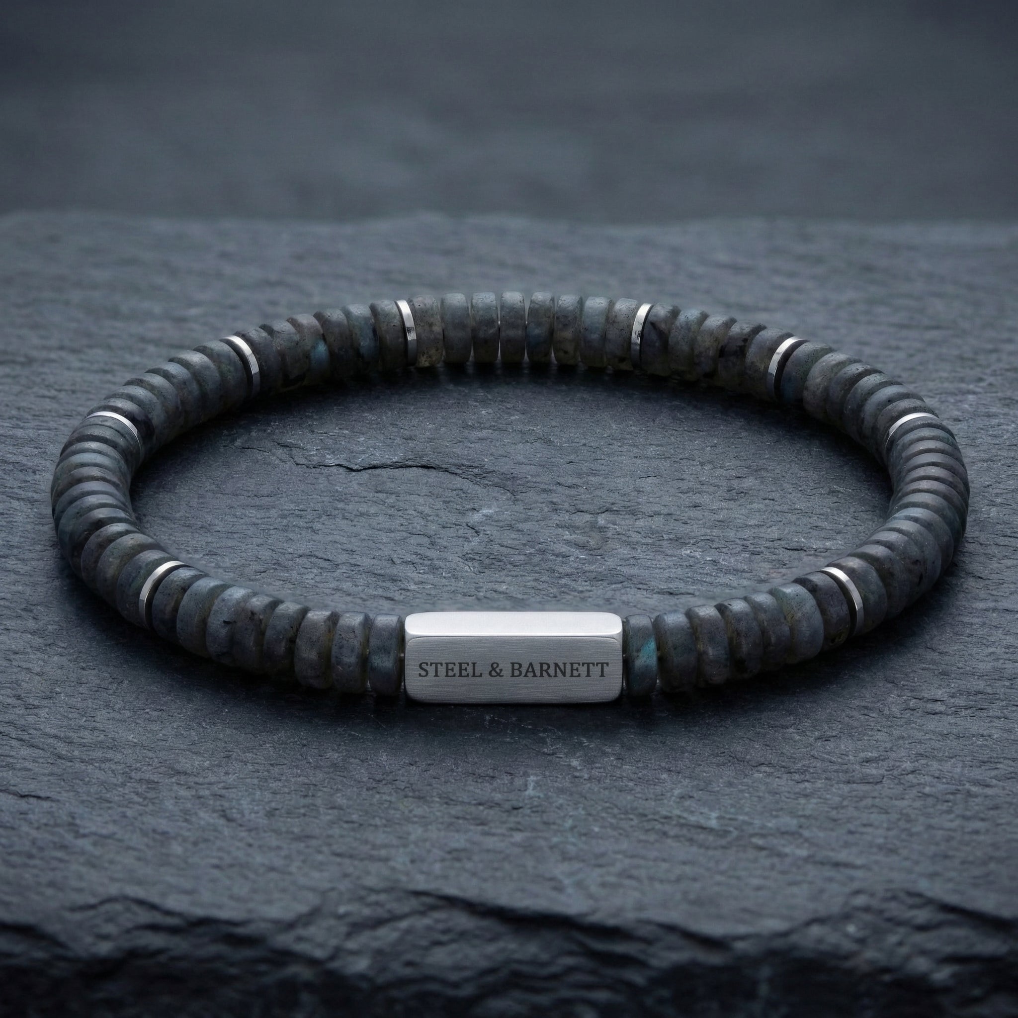 Grey natural stone men's beaded bracelet with matte disc beads and brushed stainless steel accent plate displayed on a dark stone surface close up detail