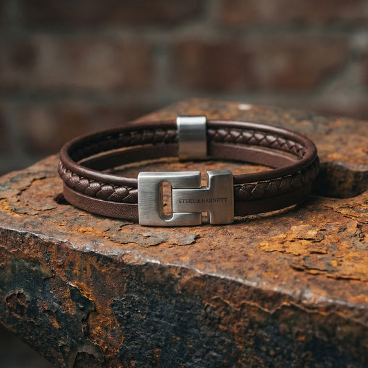 Brown multi strand leather men's bracelet with braided detail and brushed stainless steel clasp on rustic metal surface