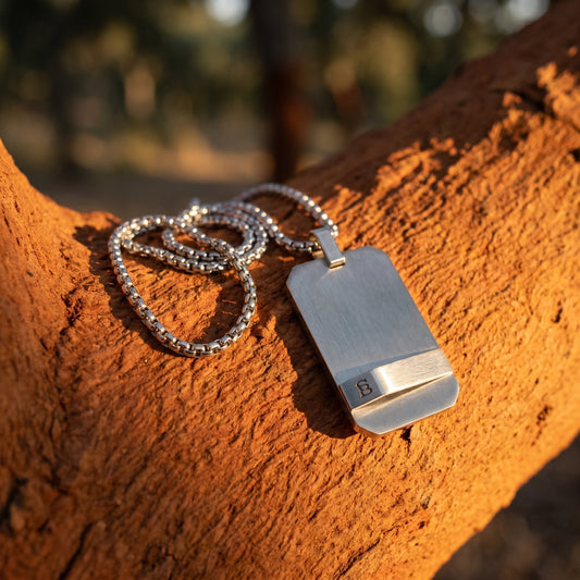 Silver rectangular pendant necklace with a brushed metal finish on a ball chain, displayed on a rustic clay surface in natural light