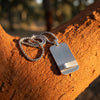 Silver rectangular pendant necklace with a brushed metal finish on a ball chain, displayed on a rustic clay surface in natural light
