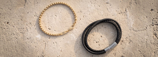 Two bracelets displayed on a textured beige stone surface: a gold beaded bracelet on the left and a black braided leather bracelet with a silver clasp on the right.