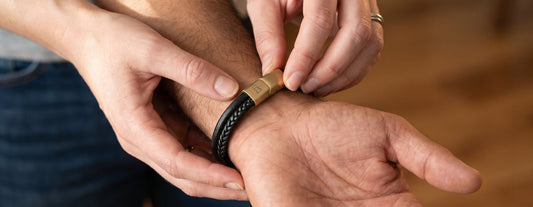 A close-up lifestyle shot of a person's hands adjusting a black braided leather bracelet with a gold-toned metal clasp onto another person's wrist.