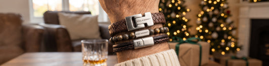 A close-up of a person's wrist wearing multiple leather and wood bracelets with silver-toned rectangular buckles, set against a cozy holiday living room background with a lit Christmas tree and fireplace.