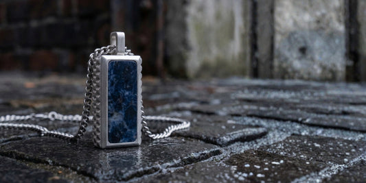 Stainless steel pendant necklace with rectangular blue natural stone and textured edges on chain, men’s jewelry displayed on wet stone pavement background