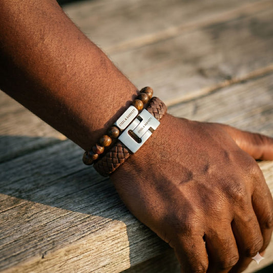 Brown braided leather men's bracelet with a brushed stainless steel clasp paired with a brown beaded bracelet, shown on wrist outdoors on a wooden surface
