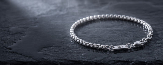 A silver beaded chain bracelet with a lobster clasp and a small engraved tag, displayed on a dark textured slate surface.
