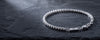 A silver beaded chain bracelet with a lobster clasp and a small engraved tag, displayed on a dark textured slate surface.