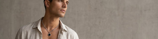 A man wearing a silver-toned necklace with a black clover-shaped pendant, styled with an open-collar light beige shirt against a textured gray concrete wall.