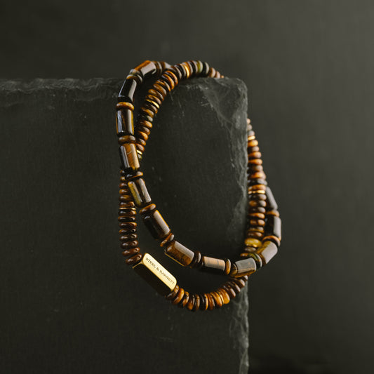 A beaded necklace with brown tiger eye and gold beads draped over a dark gray rectangular block against a dark background.
