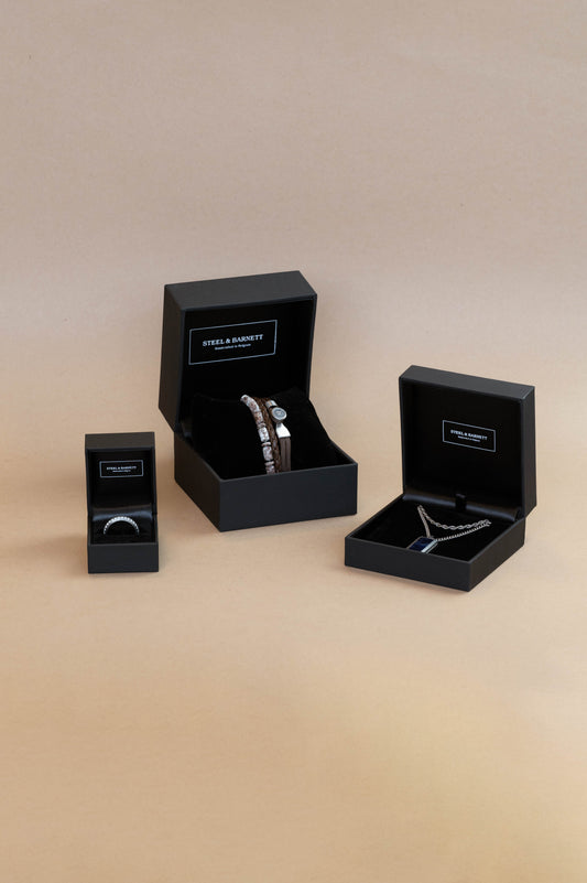 Three black jewelry gift boxes of different sizes arranged on a warm beige gradient background. Two of the boxes are open, displaying silver rings inside, and one box is closed standing to the left.
