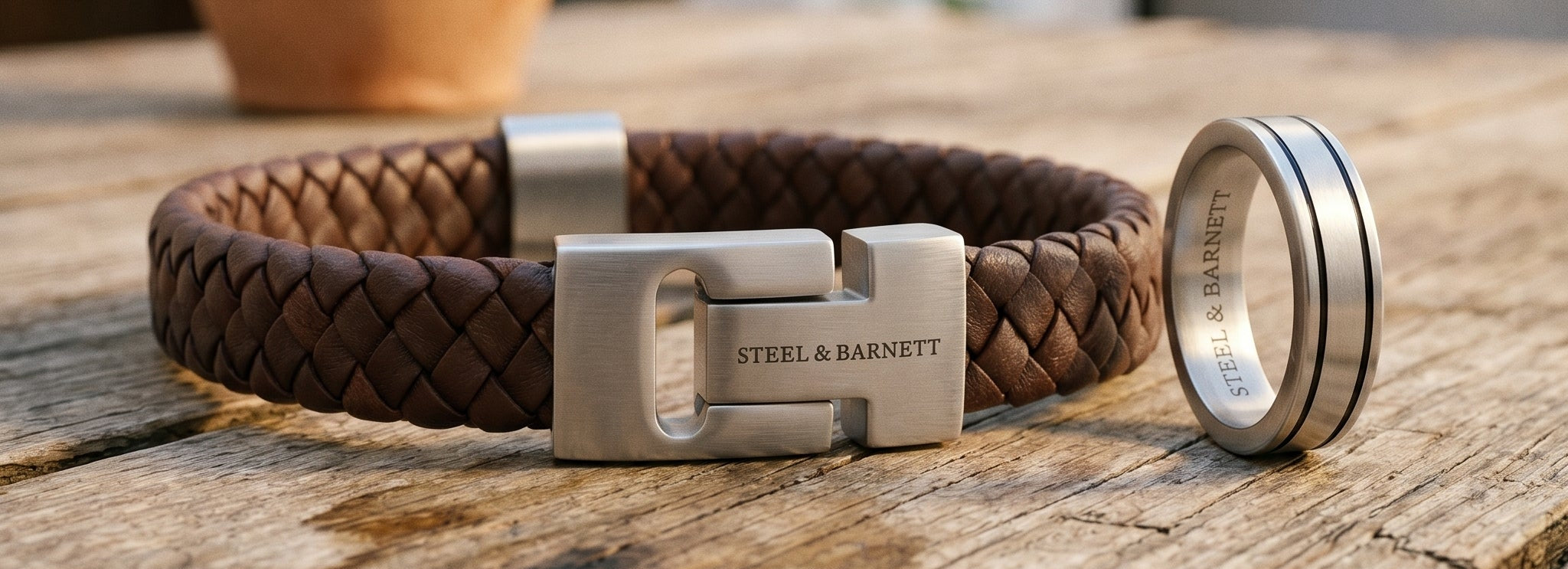 Brown braided leather men’s bracelet with brushed stainless steel clasp on a rustic wooden surface alongside a silver ring