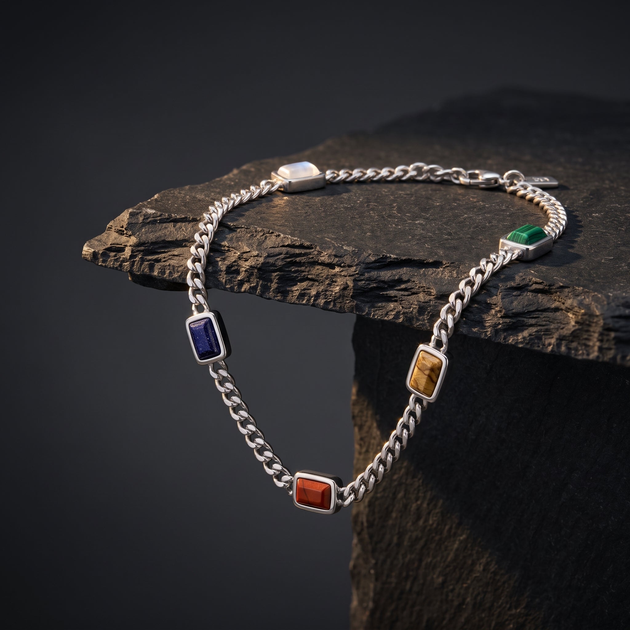 A silver-toned chain bracelet with five rectangular inlaid gemstone charms in blue, green, orange, and brown, draped over a dark textured stone block against a dark gray background.