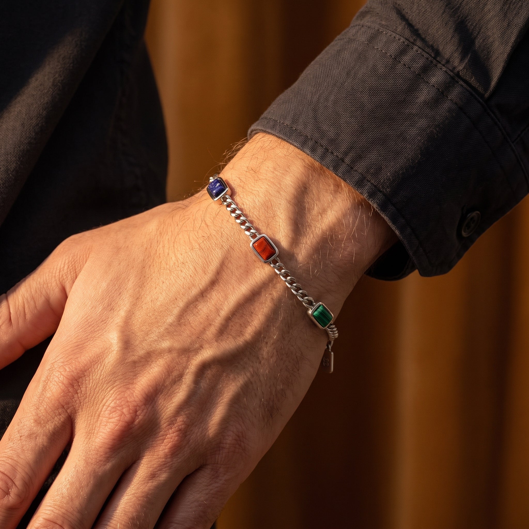 A close-up of a person's wrist wearing a silver chain bracelet with rectangular colorful stones in blue, red, and green, set against a warm brown background.