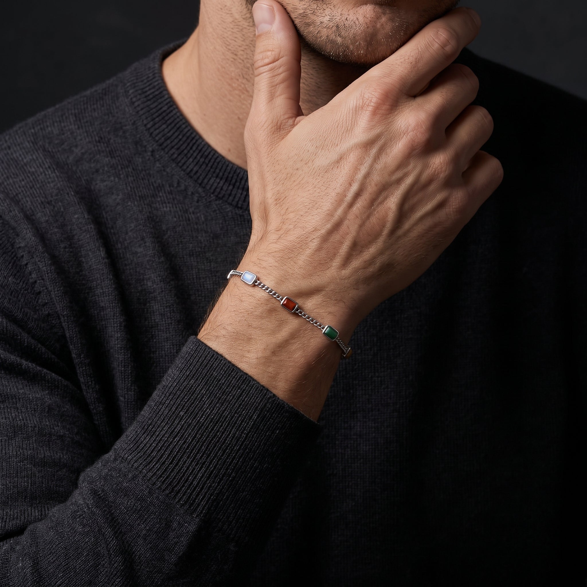 A close-up of a man wearing a silver-toned chain bracelet with colorful accents, resting his hand on his chin. He is wearing a dark charcoal gray knit sweater against a dark background.