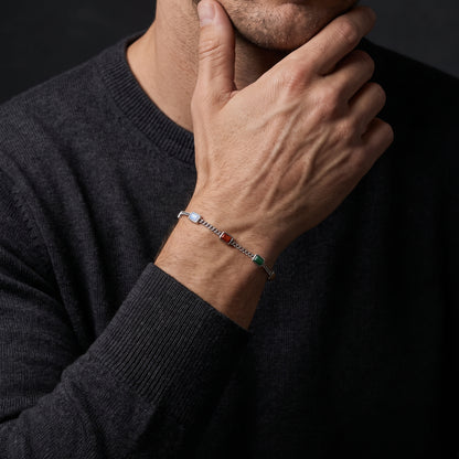 A close-up of a man wearing a silver-toned chain bracelet with colorful accents, resting his hand on his chin. He is wearing a dark charcoal gray knit sweater against a dark background.
