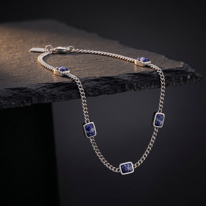 Silver chain men's bracelet with rectangular blue sodalite stone accents and a lobster clasp displayed on a dark stone surface