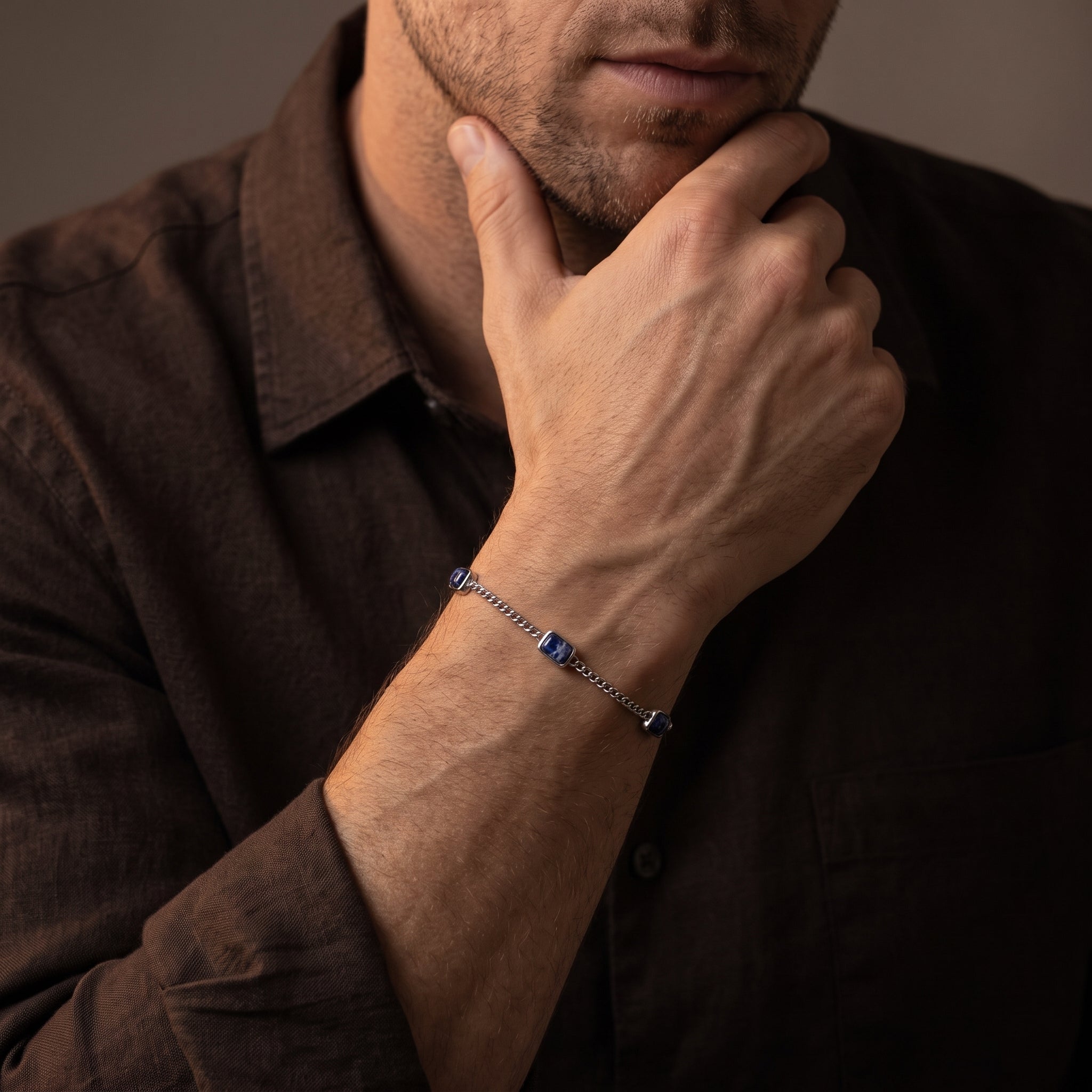 Silver chain men's bracelet with blue square stone accents worn on the wrist with a dark brown shirt