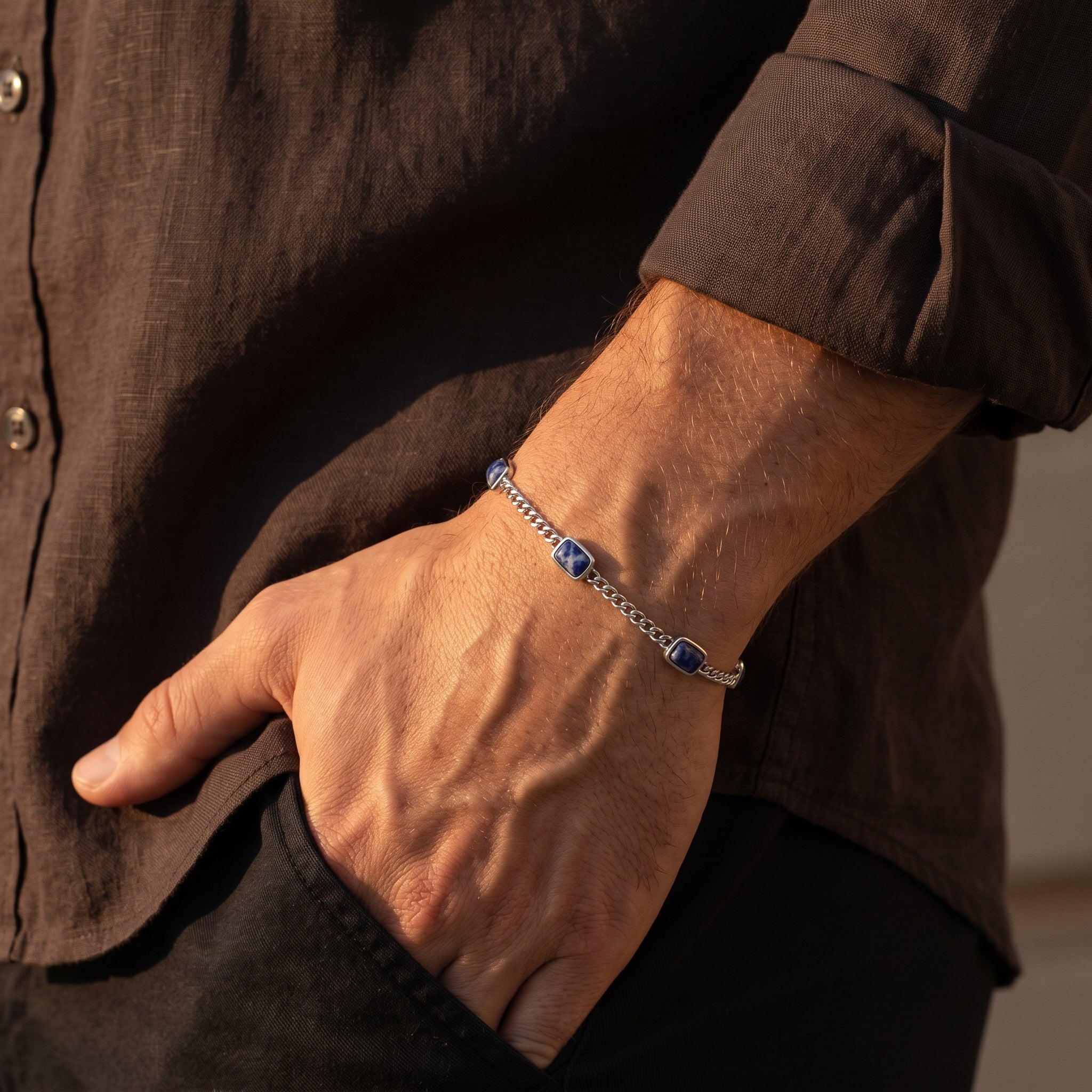 Silver chain men's bracelet with blue square stone accents worn on the wrist with a dark brown shirt