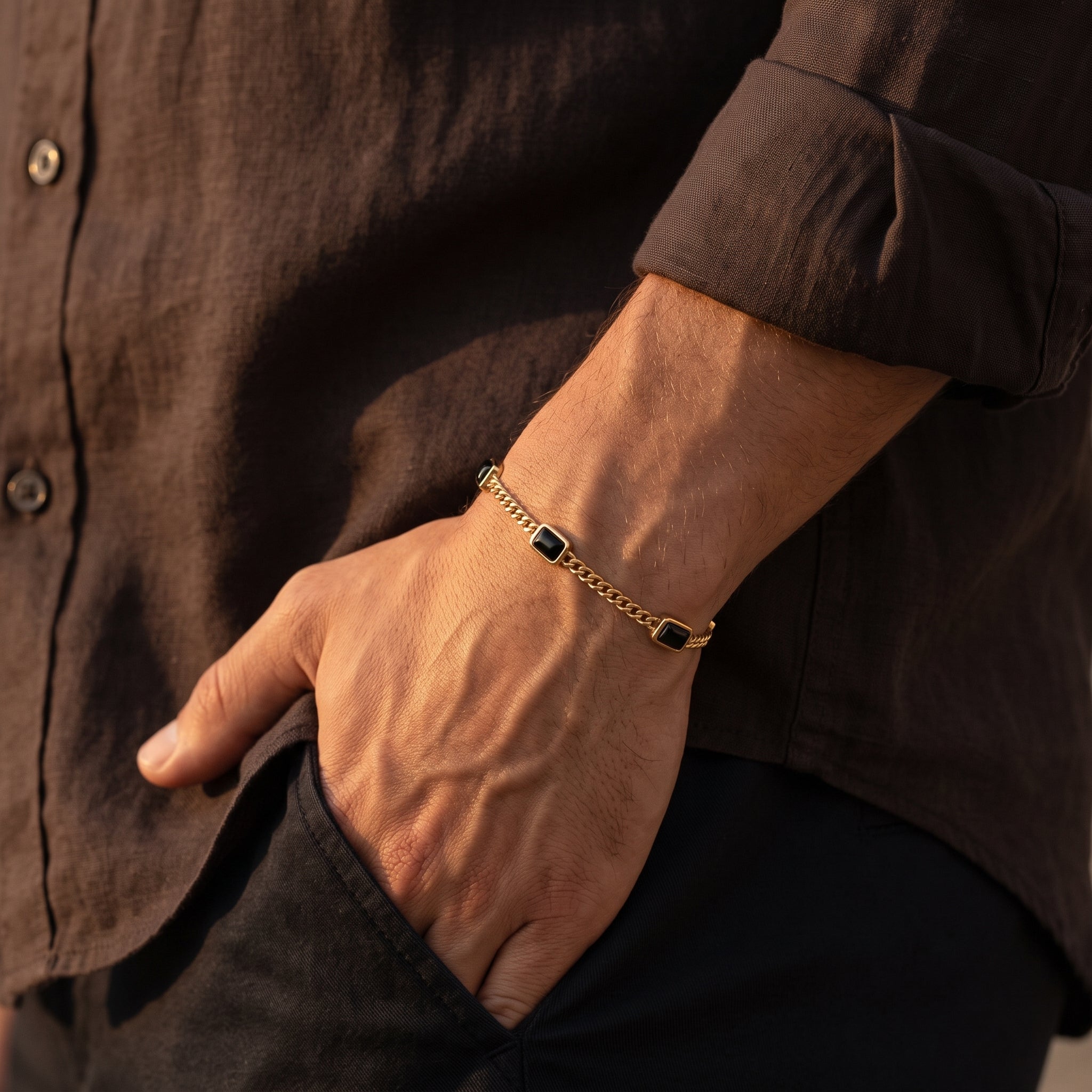 Gold chain men's bracelet with black rectangular stone accents worn on the wrist with a dark brown shirt
