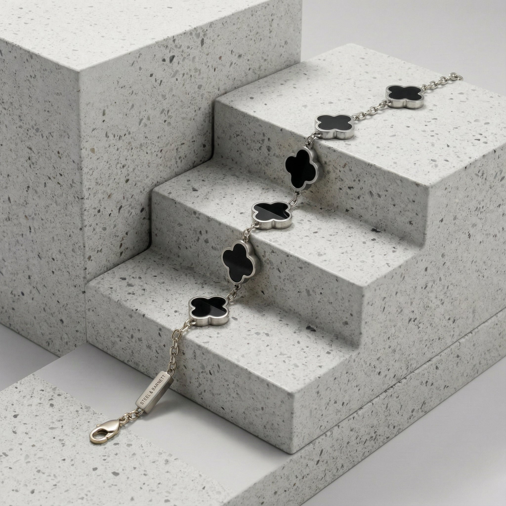 Silver toned men’s bracelet with black clover shaped inlays in stainless steel chain displayed on textured stone surface