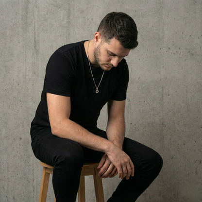 Man in black outfit with a stainless steel necklace sitting on a stool against a gray concrete wall