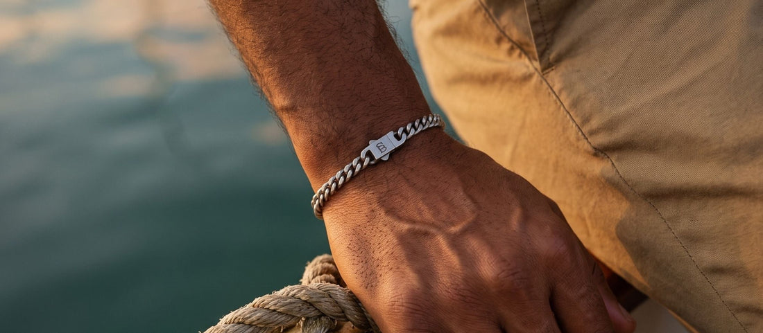A close-up lifestyle shot of a man on a boat wearing a silver chain bracelet with a lobster clasp. He is holding a wooden cleat with rope, wearing khaki pants and a cream striped shirt, with the blue-green water in the background.
