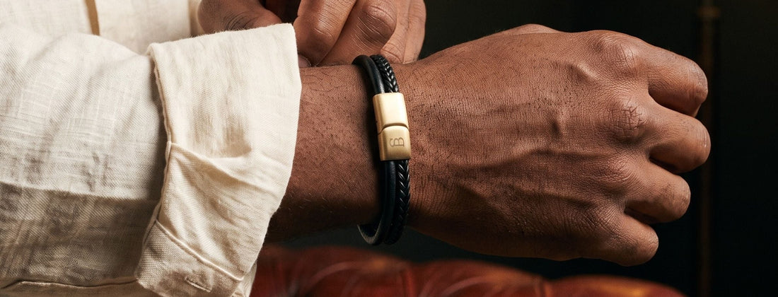 A man wearing a black braided leather bracelet with a gold-toned rectangular metal clasp engraved with 'MADE IN THE USA', shown in a lifestyle setting while adjusting the bracelet on his wrist.