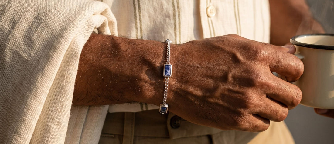 A close-up lifestyle shot of a person wearing a silver chain bracelet with a rectangular blue gemstone, holding a white enamel mug. The person is wearing a cream linen shirt and khaki trousers.