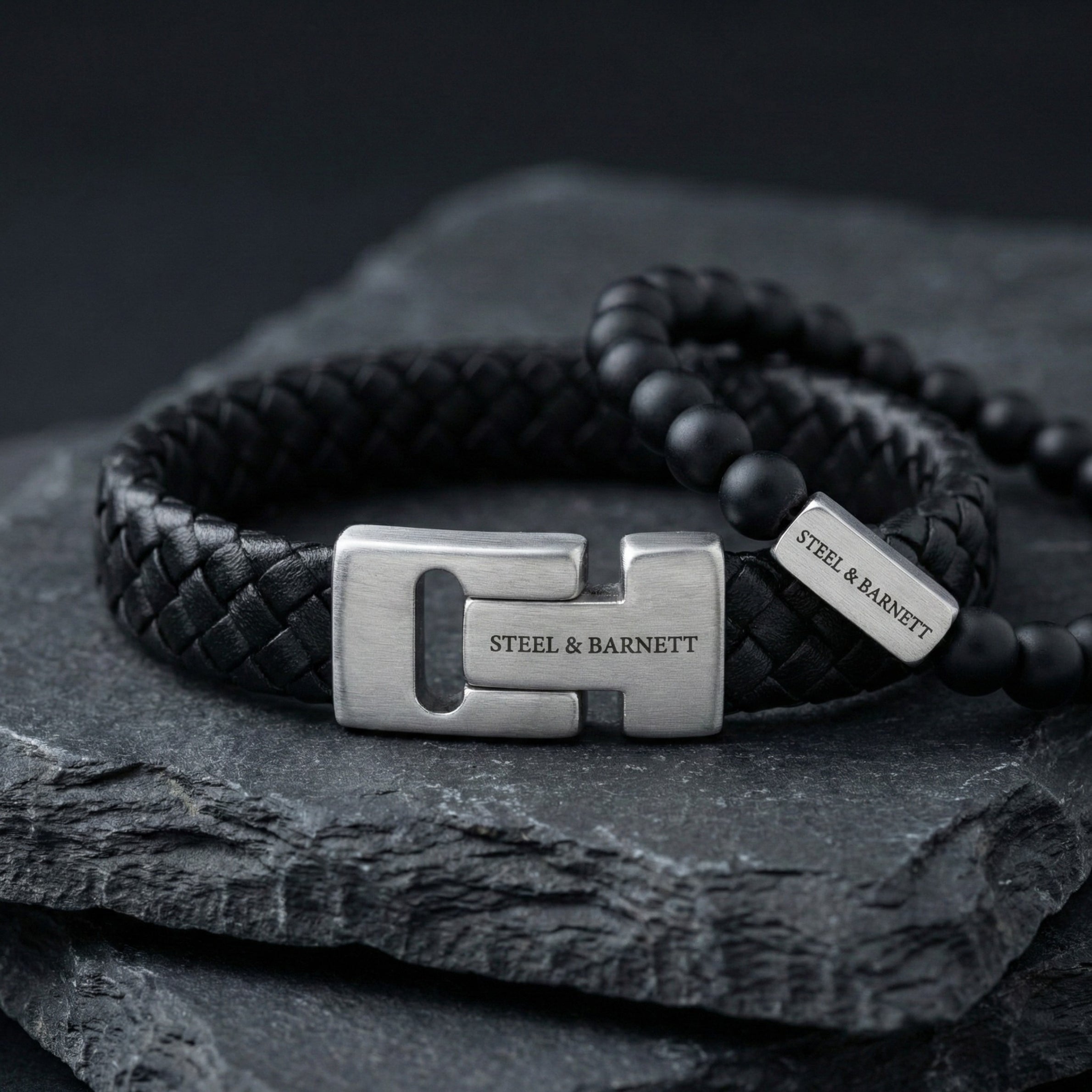 Black braided leather men's bracelet with brushed stainless steel clasp engraved Steel & Barnett shown in close up on a dark stone surface