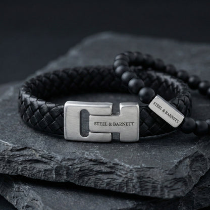 Black braided leather men's bracelet with brushed stainless steel clasp engraved Steel & Barnett shown in close up on a dark stone surface