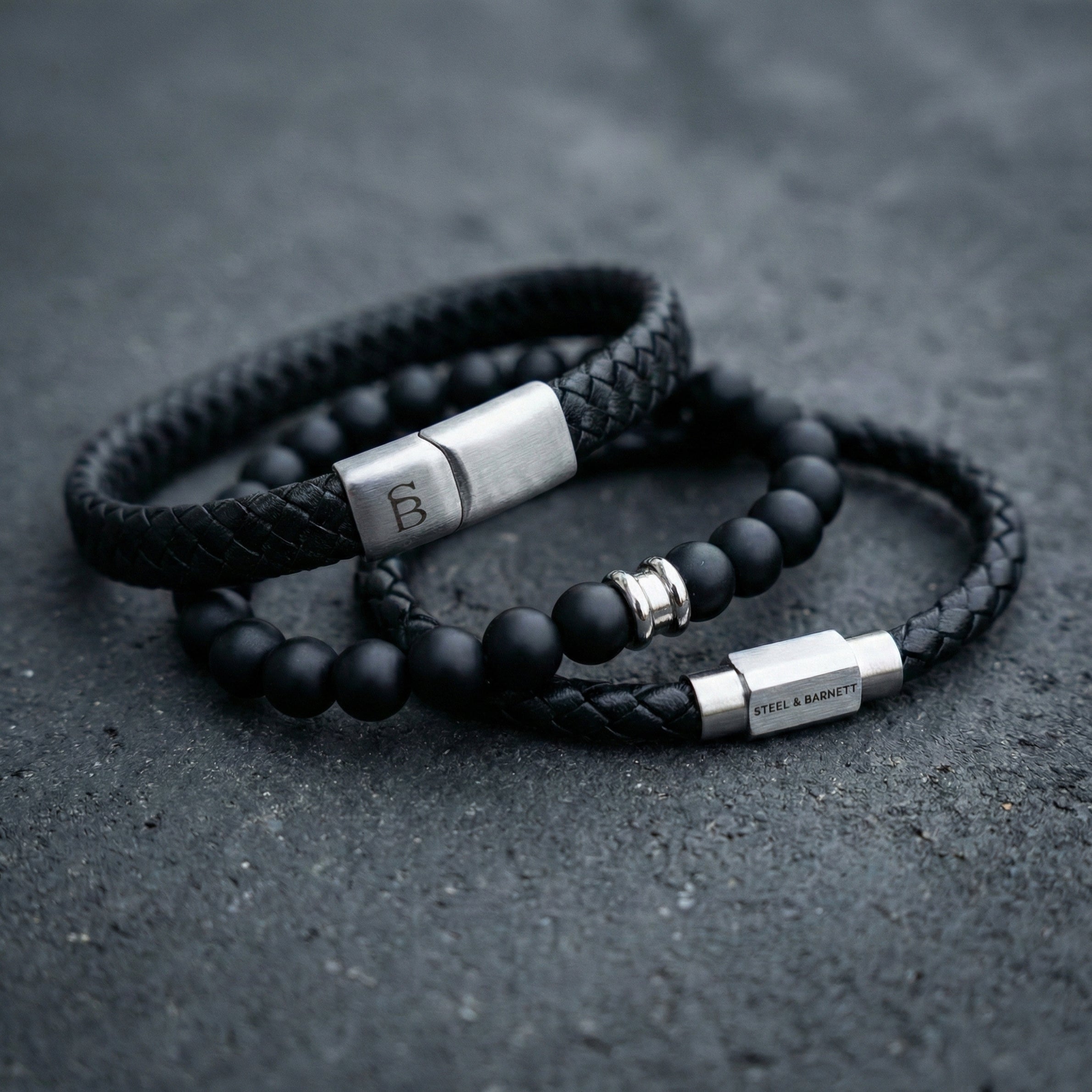 Black braided leather men’s bracelet set with matte black bead bracelet and brushed stainless steel clasp detail on dark stone surface