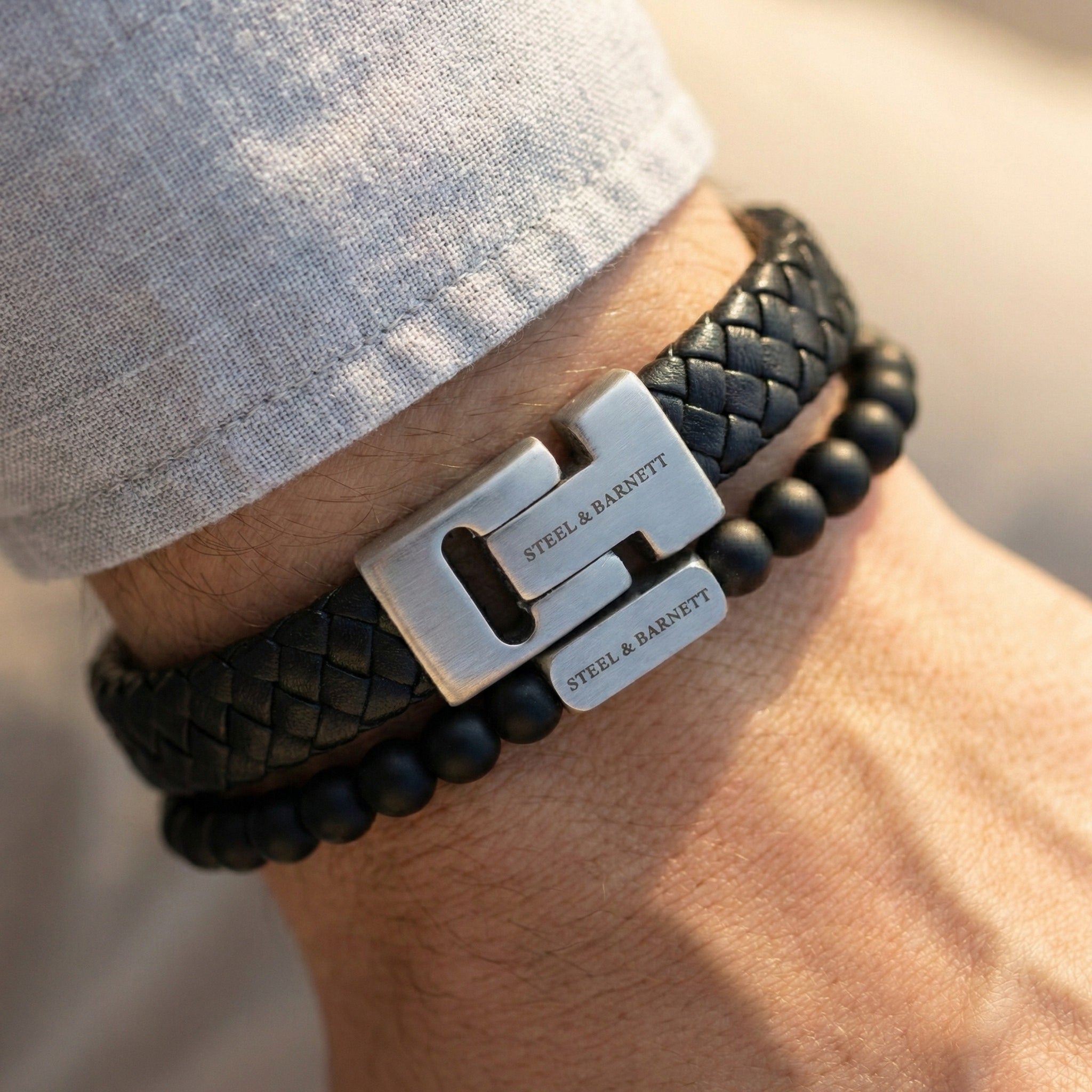 Black braided leather men's bracelet with brushed stainless steel clasp and matte black beads worn on wrist in a lifestyle setting