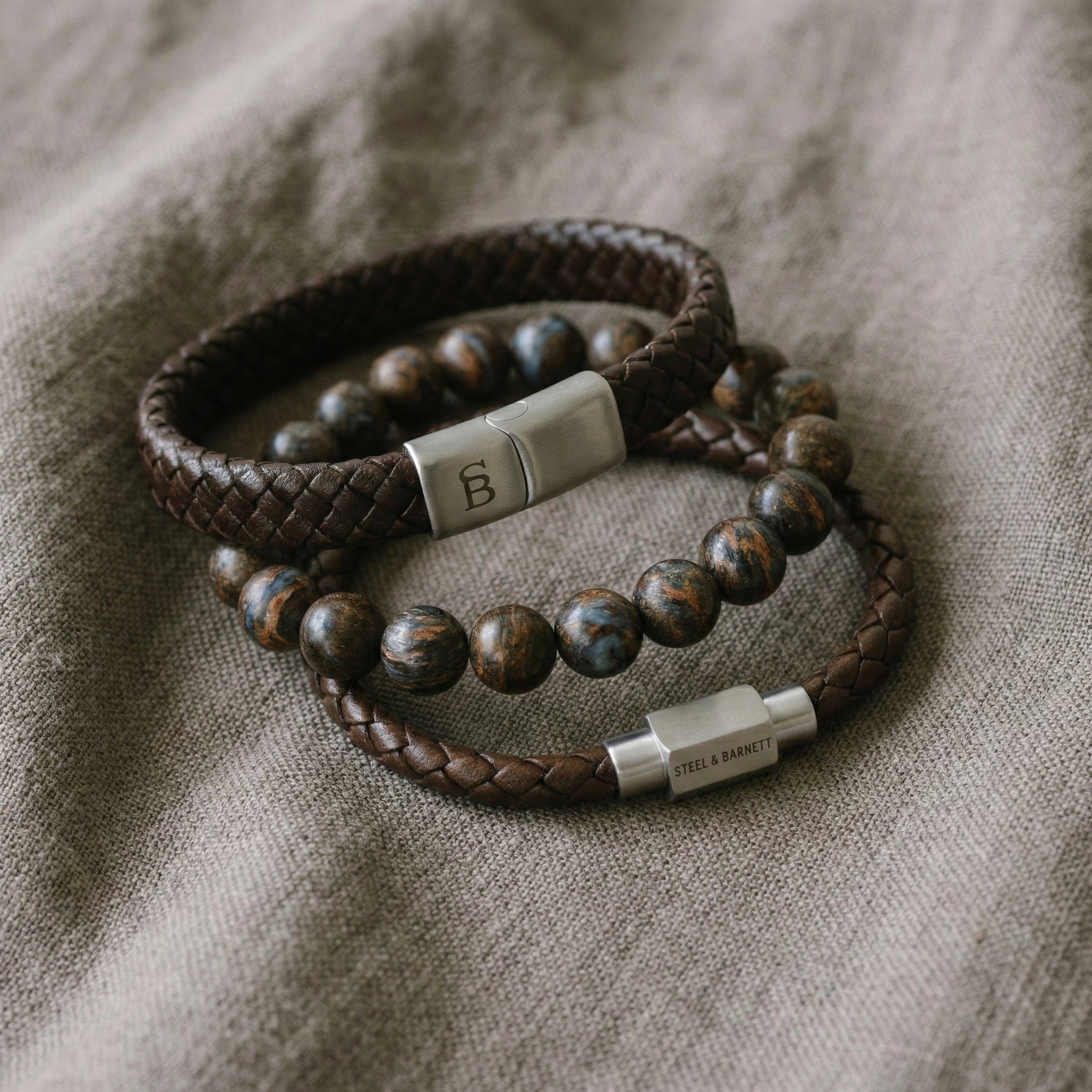 Brown braided leather men’s bracelet set with natural stone bead bracelet and brushed stainless steel clasps on beige fabric surface