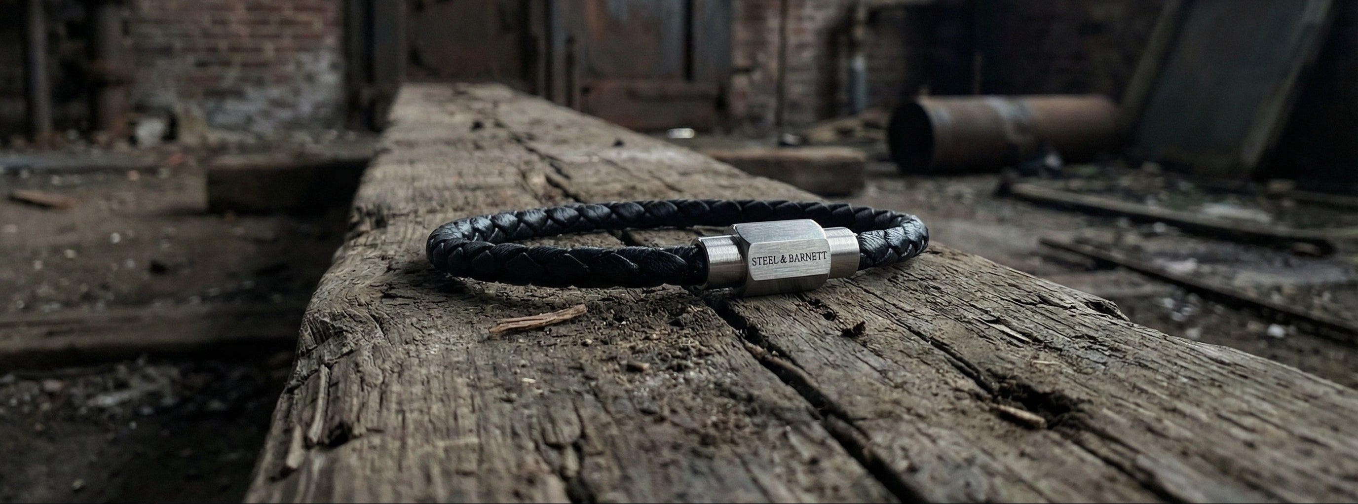 Black braided leather men's bracelet with brushed stainless steel magnetic clasp on a rustic wooden surface in an industrial setting close-up
