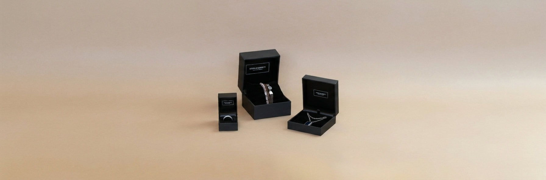 Three black jewelry gift boxes of different sizes arranged on a warm beige gradient background. Two of the boxes are open, displaying silver rings inside, and one box is closed standing to the left.