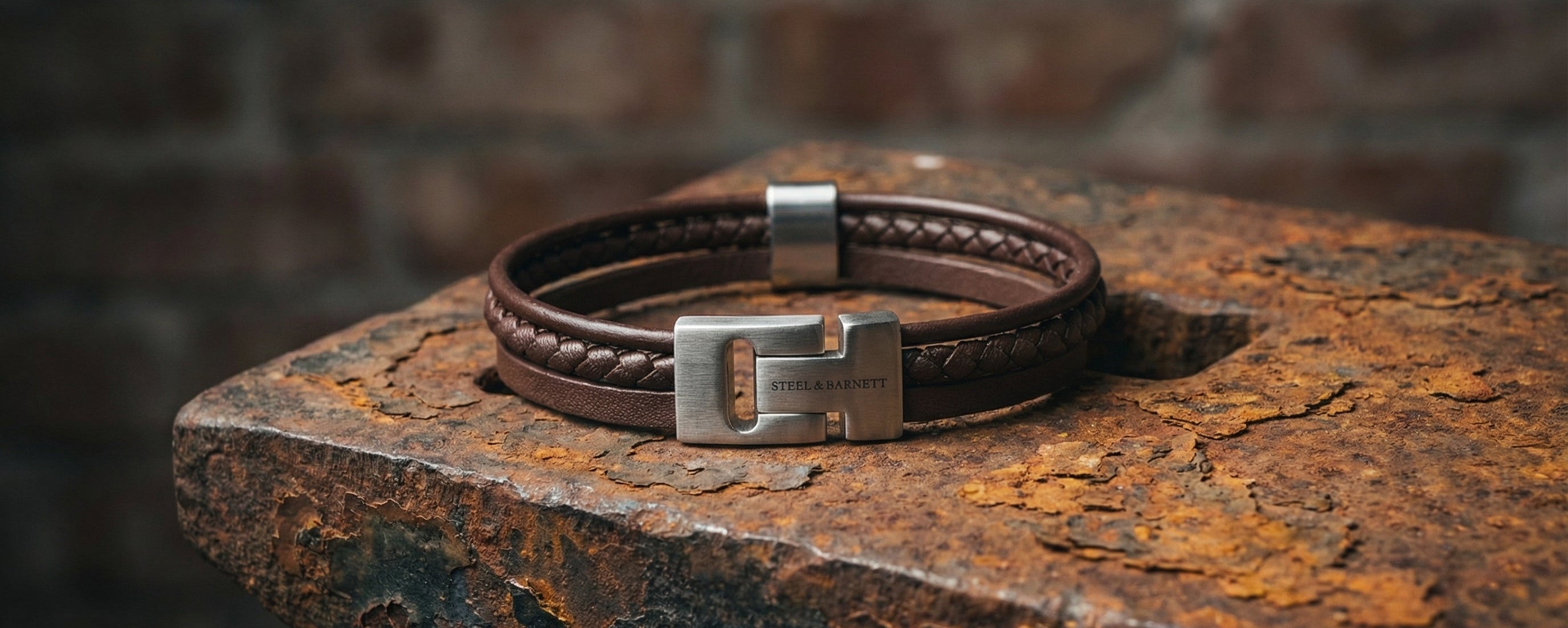 Brown leather men's bracelet with layered braided and smooth straps and brushed stainless steel hook clasp on a rusted metal surface in close-up
