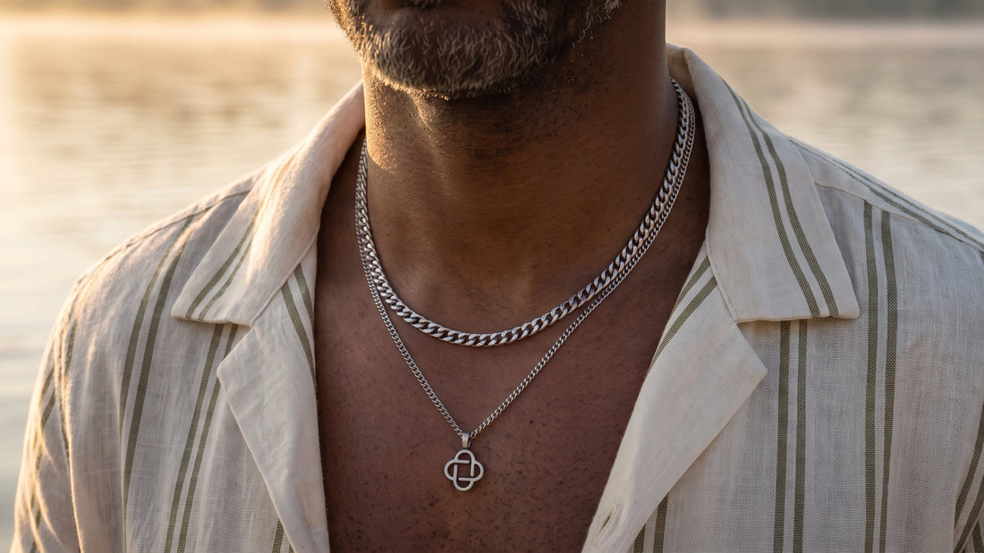 A close-up of a man wearing a silver chain necklace with a geometric pendant over a matching chain necklace, wearing an open cream-colored striped shirt against a blurred water background.