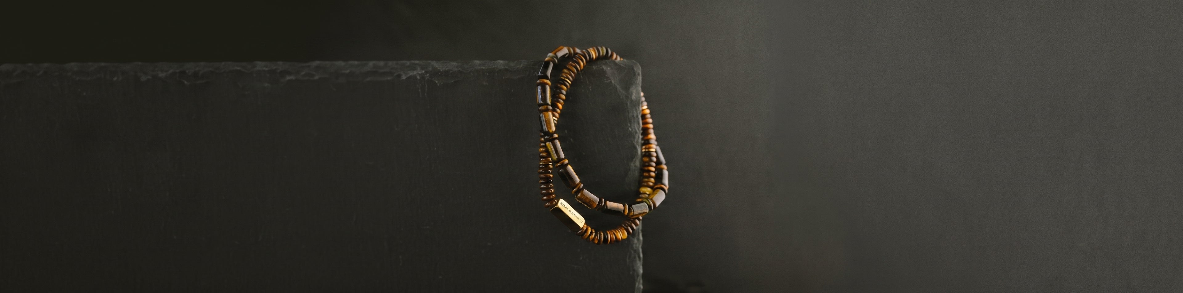 A beaded necklace with brown tiger eye and gold beads draped over a dark gray rectangular block against a dark background.