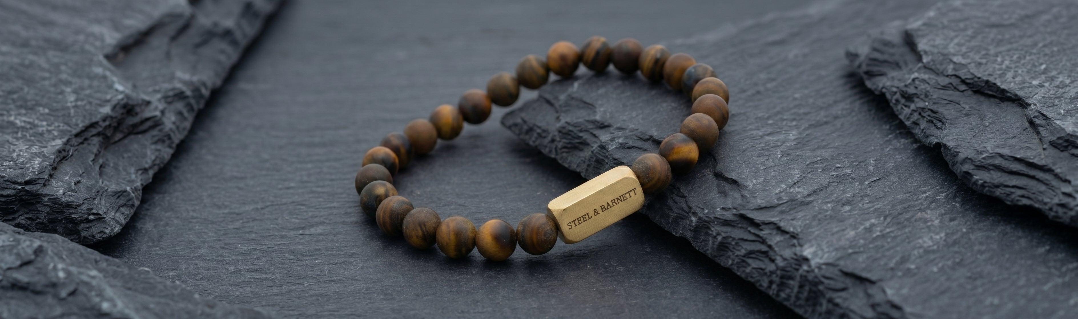 Brown tiger eye beaded men's bracelet with polished round stones and rectangular gold tone accent bead on dark slate surface